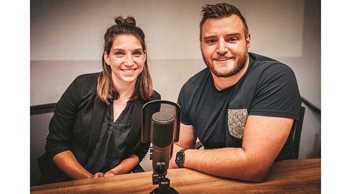 Zwei Personen sitzen an einem Tisch vor einem Mikrofon und bereiten sich auf eine Podcastaufnahme vor.