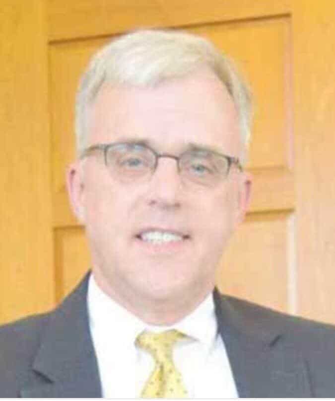 A smiling middle-aged man with graying hair and glasses wearing a suit and tie. He is facing the camera in front of a wooden door.