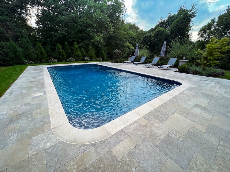 Rectangular outdoor swimming pool with clear blue water, surrounded by stone tiles, four lounge chairs, and greenery in the background.