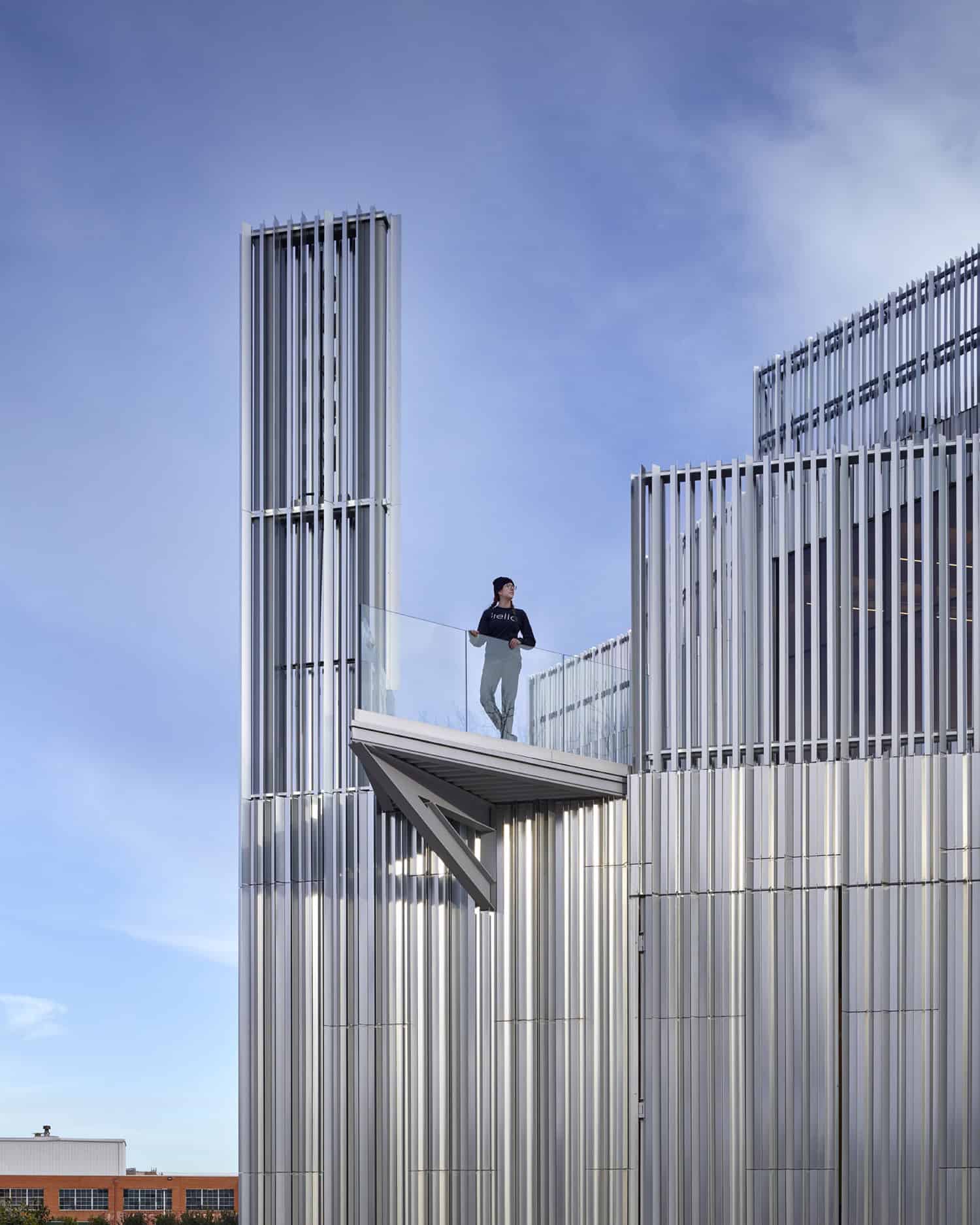 A person standing on the cantilevered balcony of the Oklahoma Contemporary Arts Center, framed by vertical aluminum fins and a blue sky.