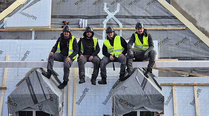 Zwei Bauarbeiter mit Warnwesten und Helmen sitzen auf einer Baustelle vor einer Wand mit Isoliermaterialien und Bauelementen.