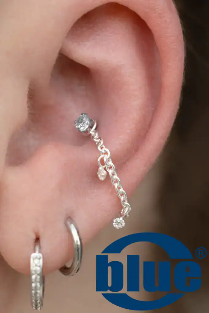 Close-up of an ear wearing a dangling diamond chain earring with a small diamond charm; blue logo watermark visible.