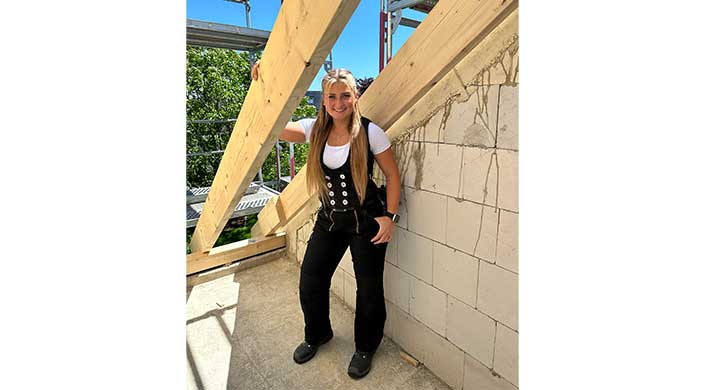 Eine Frau in traditioneller Handwerkskleidung steht auf einer Baustelle und lächelt in die Kamera.