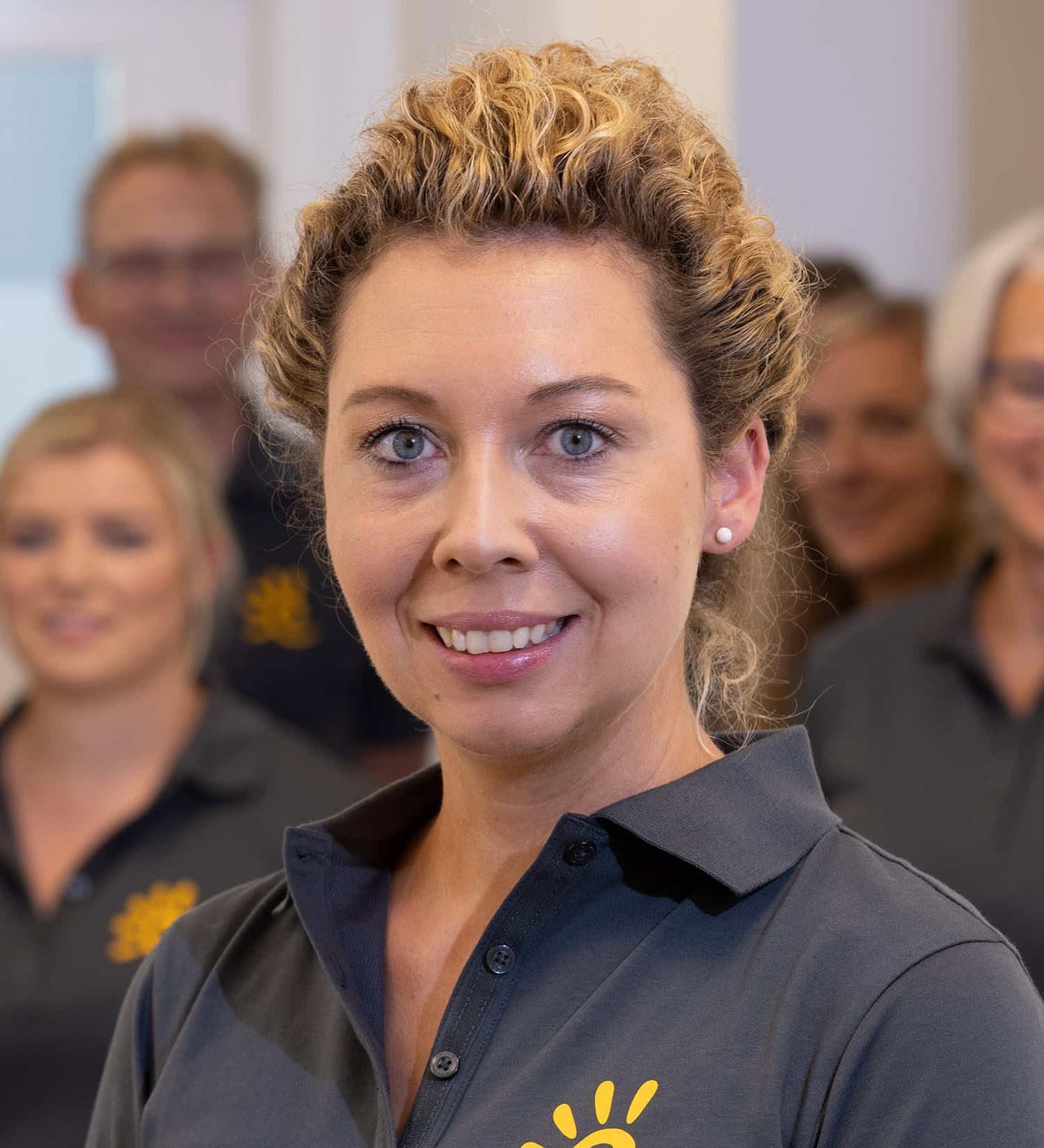 Lächelnde Mitarbeiterin in Arbeitskleidung mit dem Praxis-Team im Hintergrund.