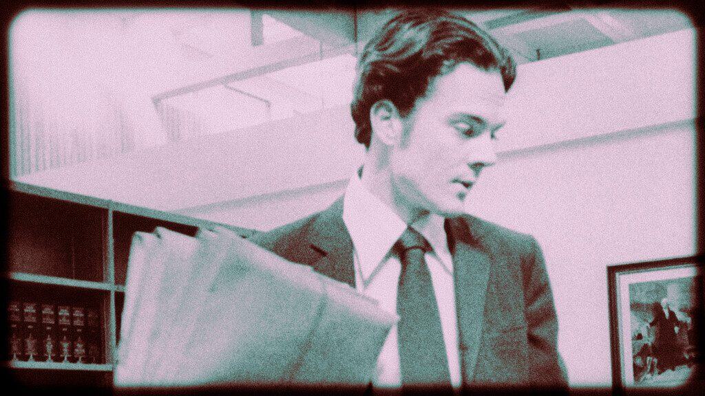 A young man in a suit reads a newspaper in a 1970s office setting.