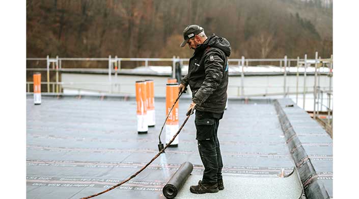 Ein Arbeiter installiert Dachbedachung mit einem Brenner, erhitzt eine Bitumenbahn auf einem Flachdach. Im Hintergrund sind weitere Rollen und Bäume sichtbar.