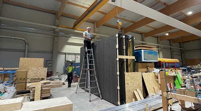 Ein Mann steht auf einer Leiter und arbeitet an einer großen, dunklen Holzwand in einer Werkhalle mit sichtbaren Holzbalken.