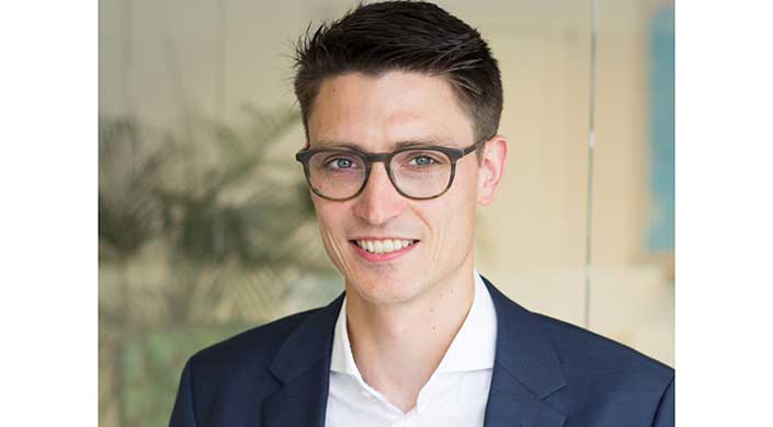 Ein Mann mit braunen Haaren, Brille und Anzug lächelt in die Kamera.