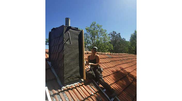 Eine Person sitzt auf einem schrägen Ziegeldach neben einem Dachfenster, trägt Arbeitskleidung und einen Hut. Im Hintergrund sind Bäume und blauer Himmel zu sehen.