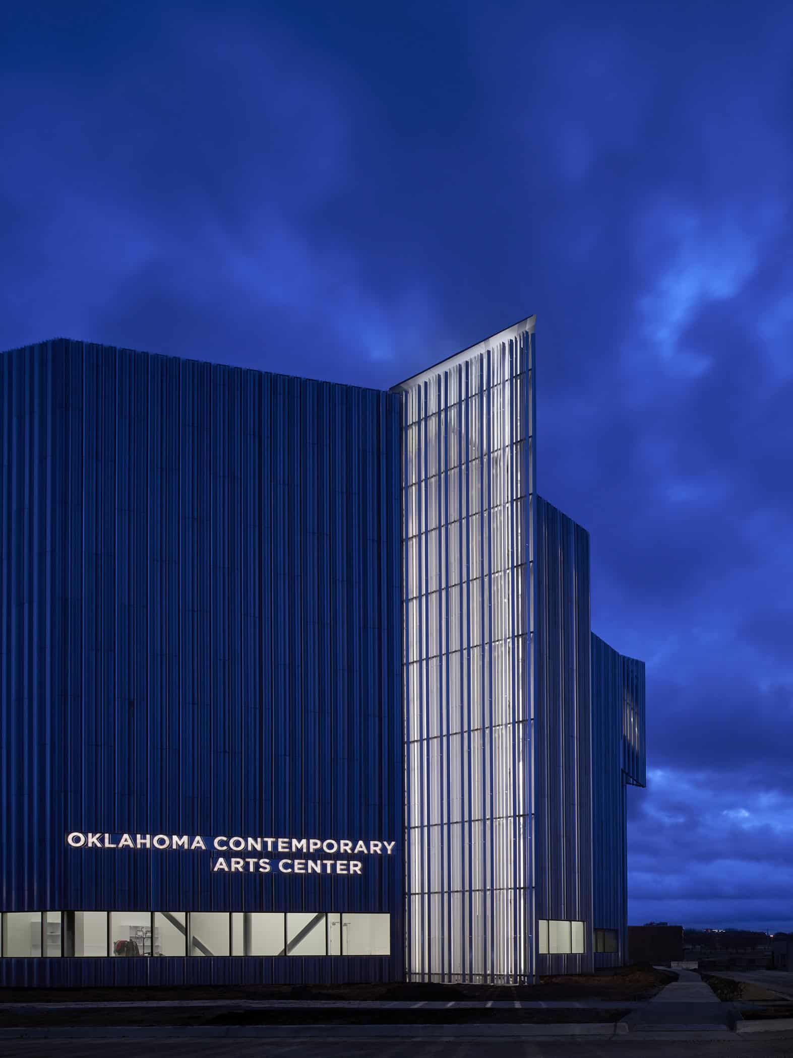 Exterior view of the Oklahoma Contemporary Arts Center at dusk, showcasing the illuminated vertical aluminum fins and architectural design.