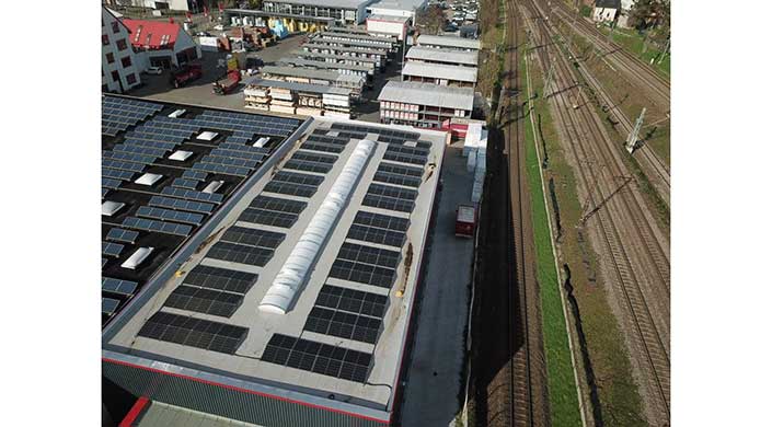 Großes Fabrikdach mit mehreren Solarpanelen, umgeben von weiteren Industriegebäuden, Blick aus der Vogelperspektive.