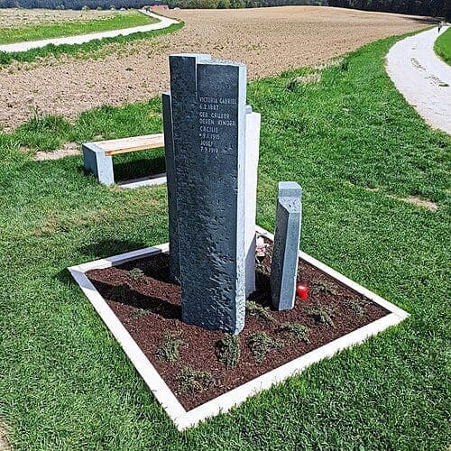 A memorial of tall grey stone slabs engraved with names, set in a bed of soil with a low white border, beside a bench and a path between fields