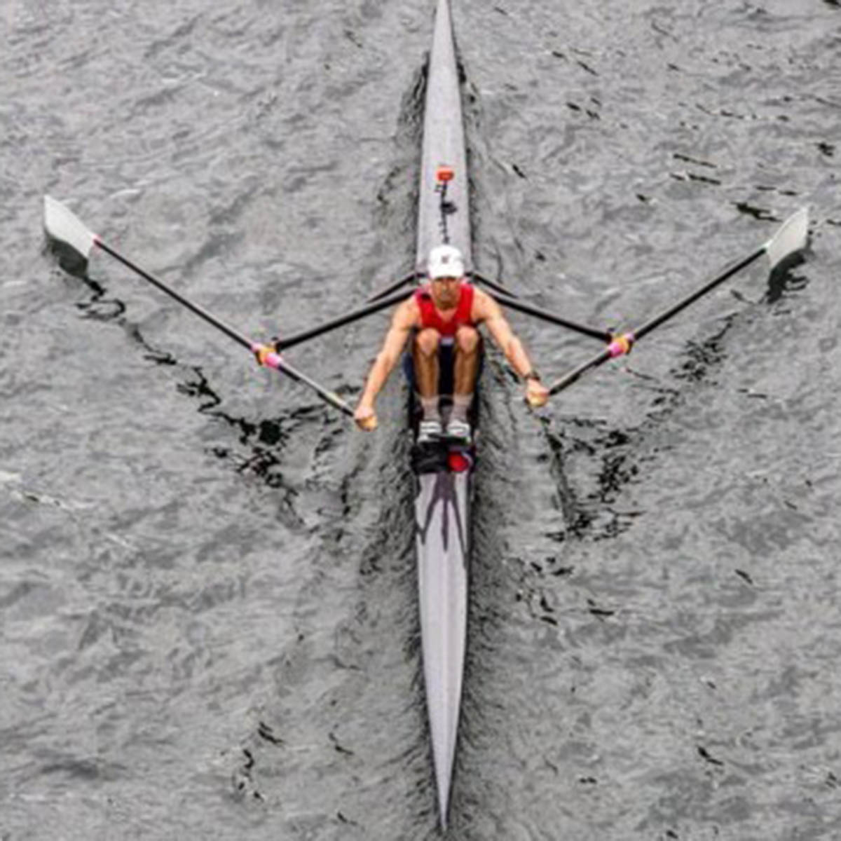 A single rower wearing a helmet and red top rows a narrow racing shell on rippled water, viewed from above.