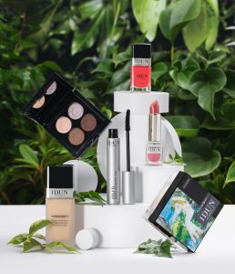 Een compositie van IDUN Minerals make-upproducten tentoongesteld op witte, cirkelvormige blokken tegen een achtergrond van groene bladeren. De collectie bevat een foundationfles, een open oogschaduwpalette met neutrale tinten, een mascara met een zilveren huls, een rode nagellak, een roze lipstick en een compact poeder met een artistieke verpakking. De natuurlijke elementen en frisse kleuren benadrukken de pure en minerale samenstelling van de producten.