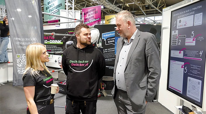 Zwei Männer unterhalten sich auf einer Messe, im Hintergrund bunte Schilder und Informationsstände.