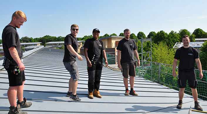 Ein Mann in Arbeitskleidung und Mütze steht auf einem Metalldach neben einer weiteren Person bei sonnigem Wetter.