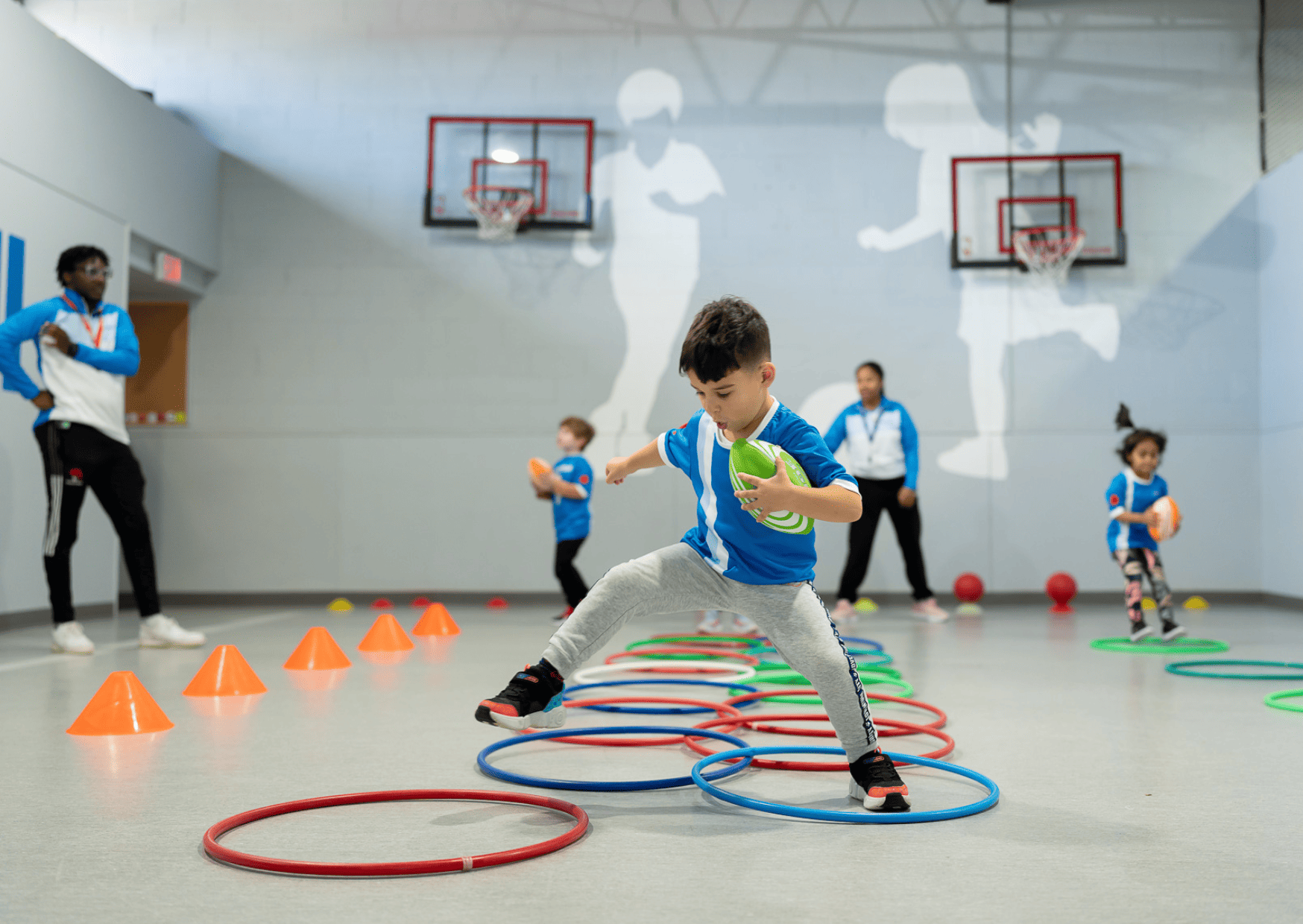 Child in a Sportball class steps through hoops while holding a ball during an indoor skills session with coaches and other children.