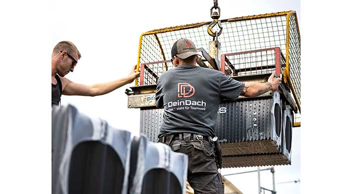 Ein Arbeiter mit "DeinDach"-T-Shirt steht vor einem Kran und trägt eine Kappe, rechteckige Metallstruktur im Hintergrund.
