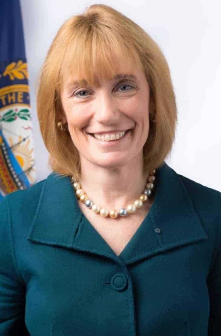 Vermont Governor Phil Scott smiles, wearing a teal blazer and a pearl necklace, standing in front of the Vermont state flag. She is a proponent of renewable energy.