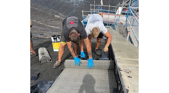 Zwei Männer mit blauen Handschuhen arbeiten konzentriert auf einer Baustelle, bücken sich über eine Kante, Baugerüst im Hintergrund, Schatten einer dritten Person sichtbar.
