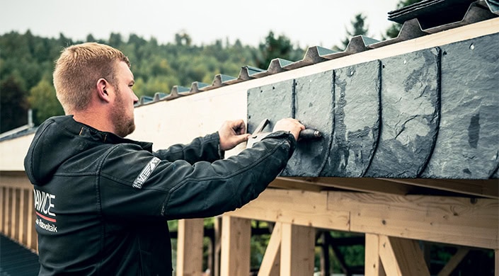 Eine Person arbeitet mit Werkzeugen an einem Dach, befestigt Dachplatten aus Schiefer auf einer Holzkonstruktion. Im Hintergrund sind Bäume und ein bewölkter Himmel zu sehen.