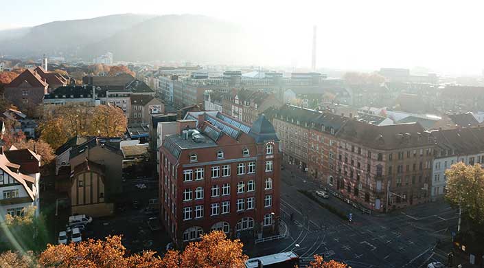 Stadtansicht mit alten Backsteingebäuden, mehreren Fensterreihen und Straßen, leichter Nebel im Hintergrund, Herbstbäume im Vordergrund.