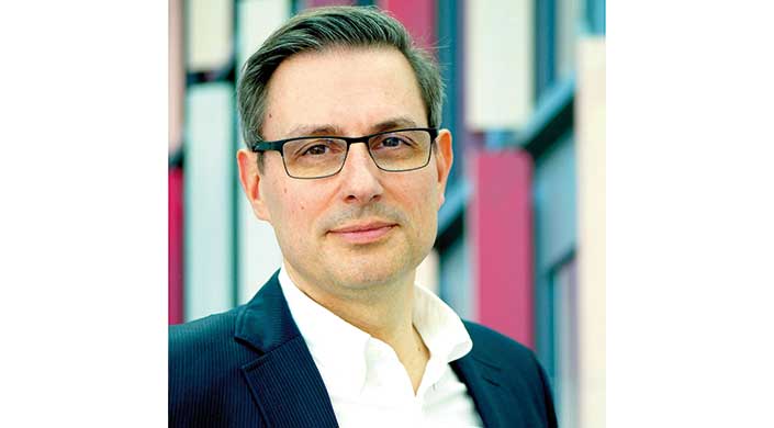 Porträt eines Mannes mit Brille, kurzem Haar, hellem Hemd und dunklem Jackett, vor unscharfem Hintergrund.