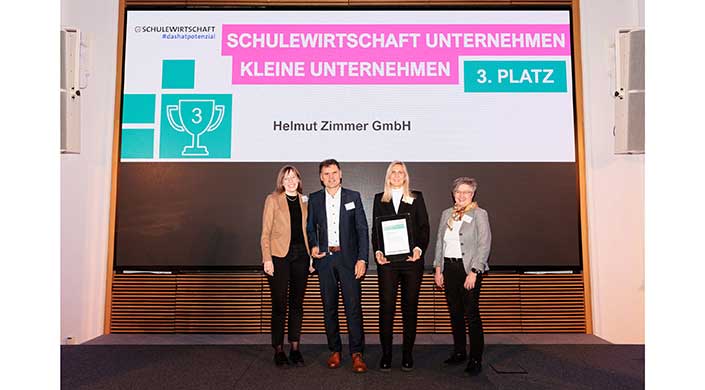 Drei Personen stehen vor einer Leinwand mit Text, zwei halten Urkunden. Sie tragen formelle Kleidung und lächeln in die Kamera.