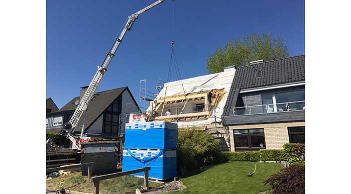 Ein Kran hebt Baumaterialien zu einem teilweise gebauten Haus, während Arbeiter auf Gerüsten stehen. Ein Stapel blauer Baumaterialien steht im Vordergrund.