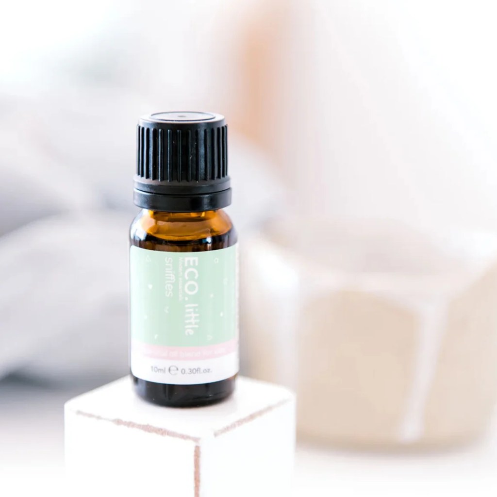 A 10ml (0.30 fl oz) amber bottle of ECO Little Sniffles Essential Oil Blend rests on a white block, with a blurred beige cup in the soft background.