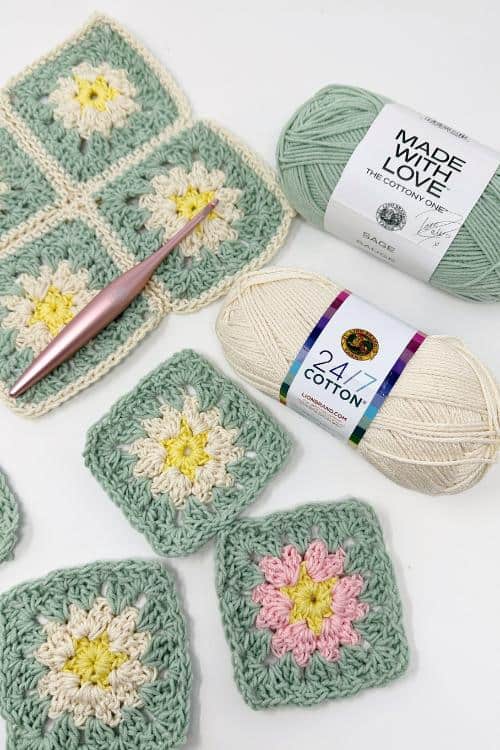 Crocheted granny squares in pastel colors, a pink crochet hook, and two skeins of cotton yarn—one mint green and one cream—are arranged on a white surface, perfect for kitchen decor or trying out a free crochet pattern.