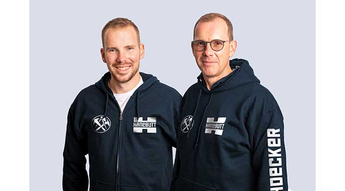Zwei Männer tragen dunkelblaue Hoodies mit Logo und Schriftzug, stehen nebeneinander vor hellem Hintergrund.