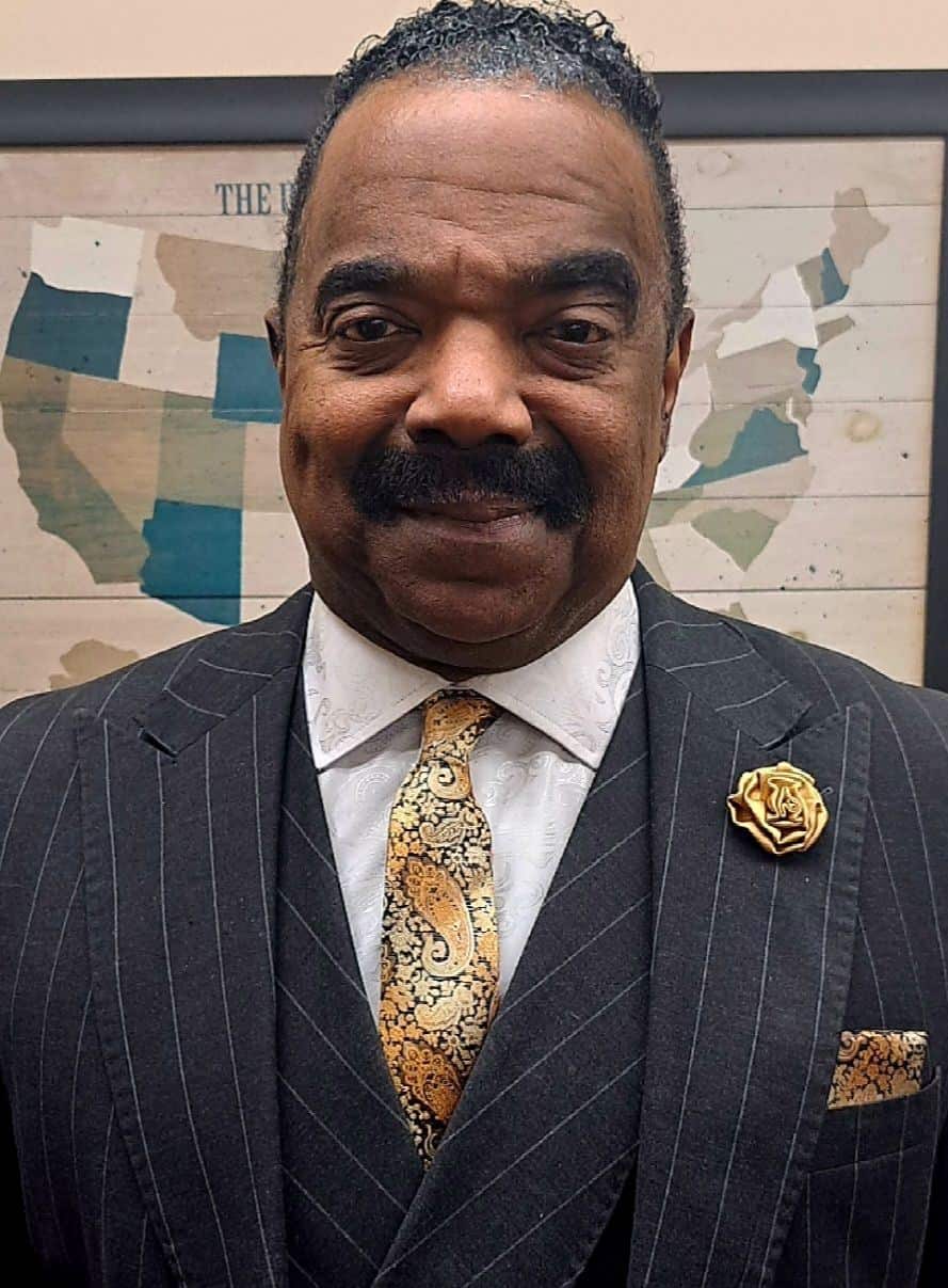 Rafael Morton Headshot A man in a pinstripe suit standing in front of The History Museum map.