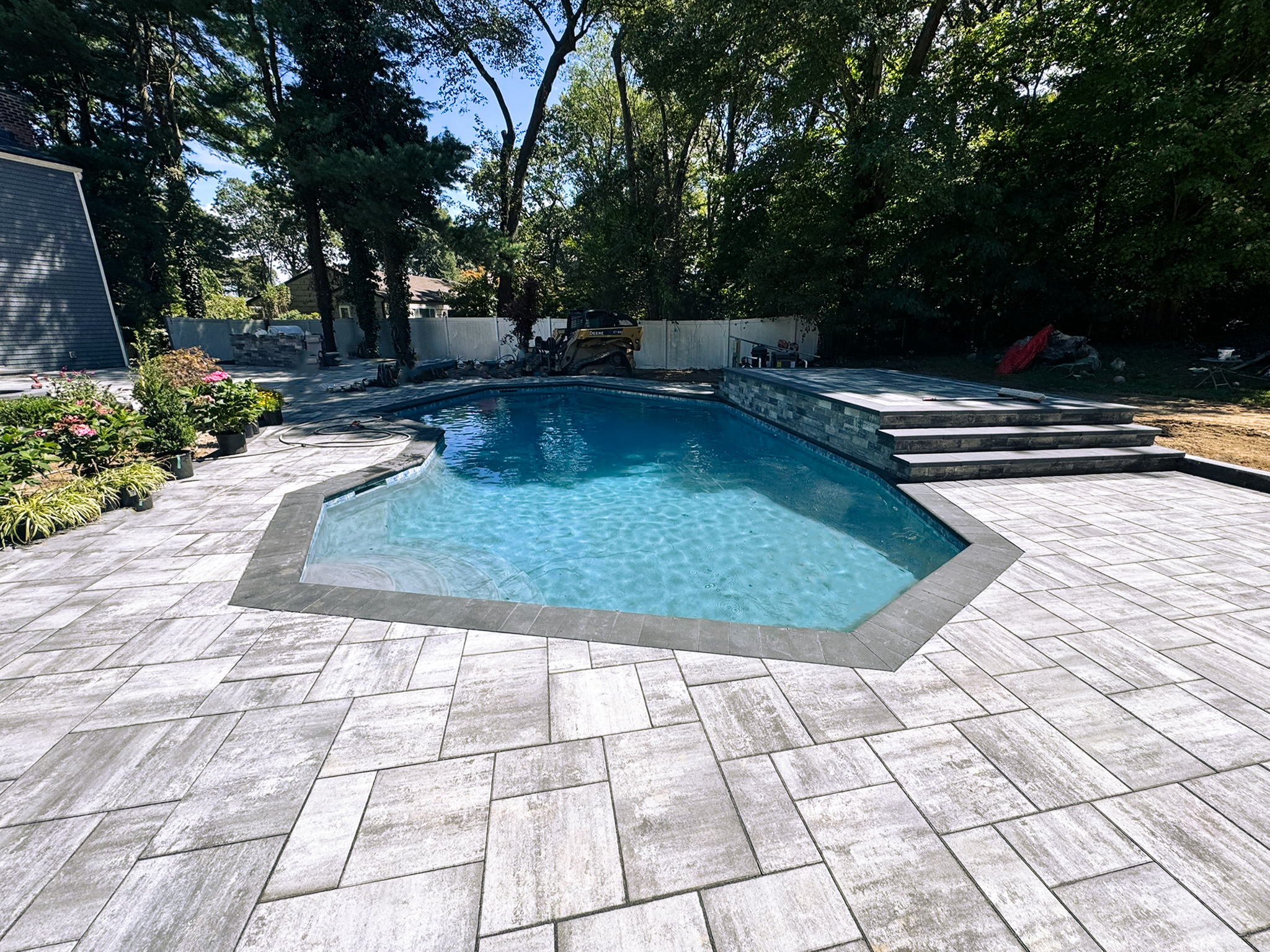 A backyard swimming pool with light gray stone pavers, surrounded by trees and landscaping, under a clear, sunny sky.