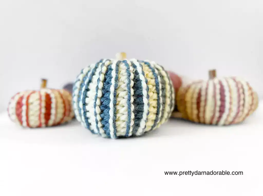 Four small chunky ribbed crochet pumpkins in multicolored crochet yarn sit on white; front has blue, cream, yellow stripes. Website promo text in corner. Free crochet pattern