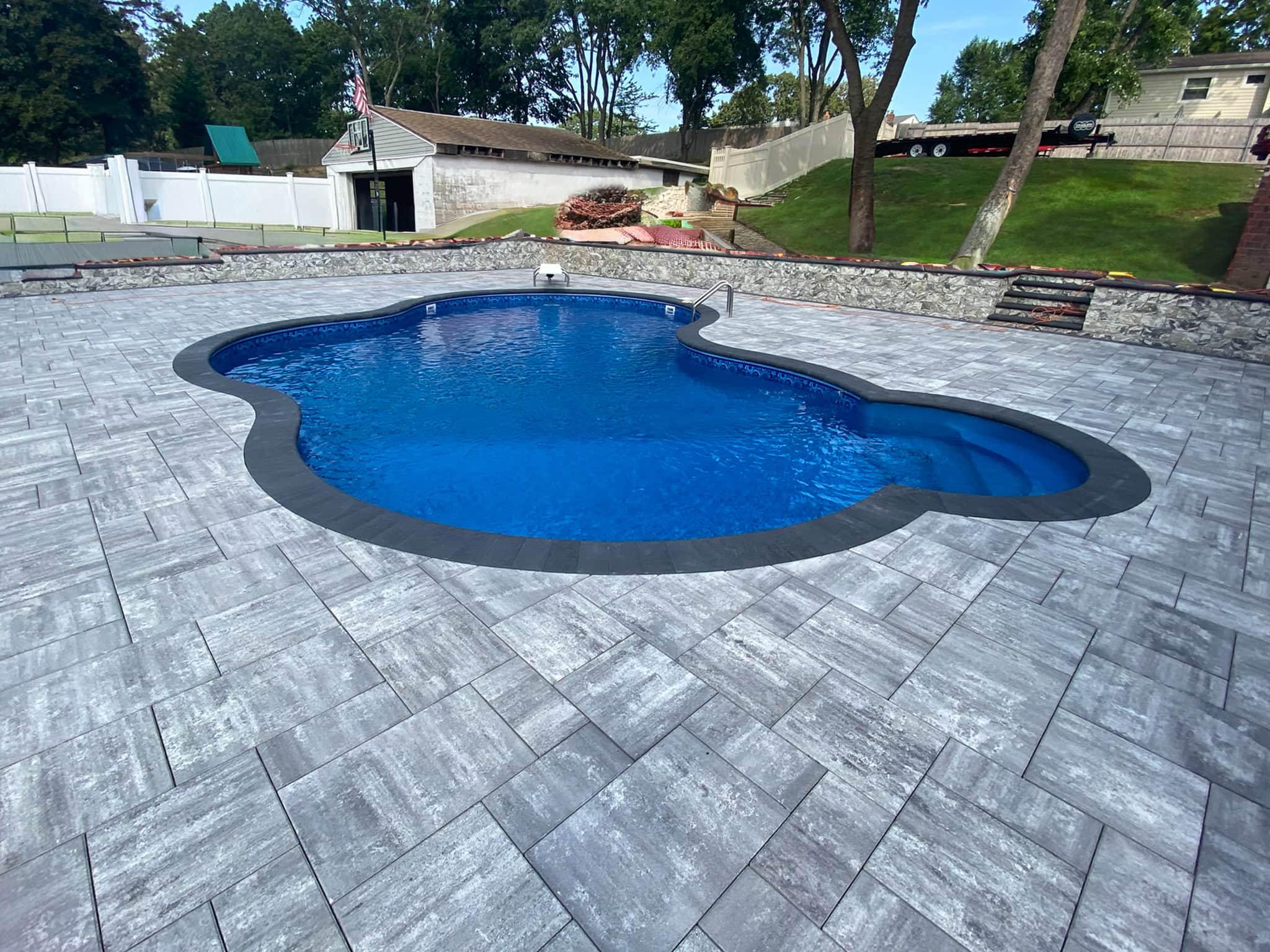 A kidney-shaped in-ground swimming pool is surrounded by gray stone pavers in a backyard with trees, a white fence, and a shed.