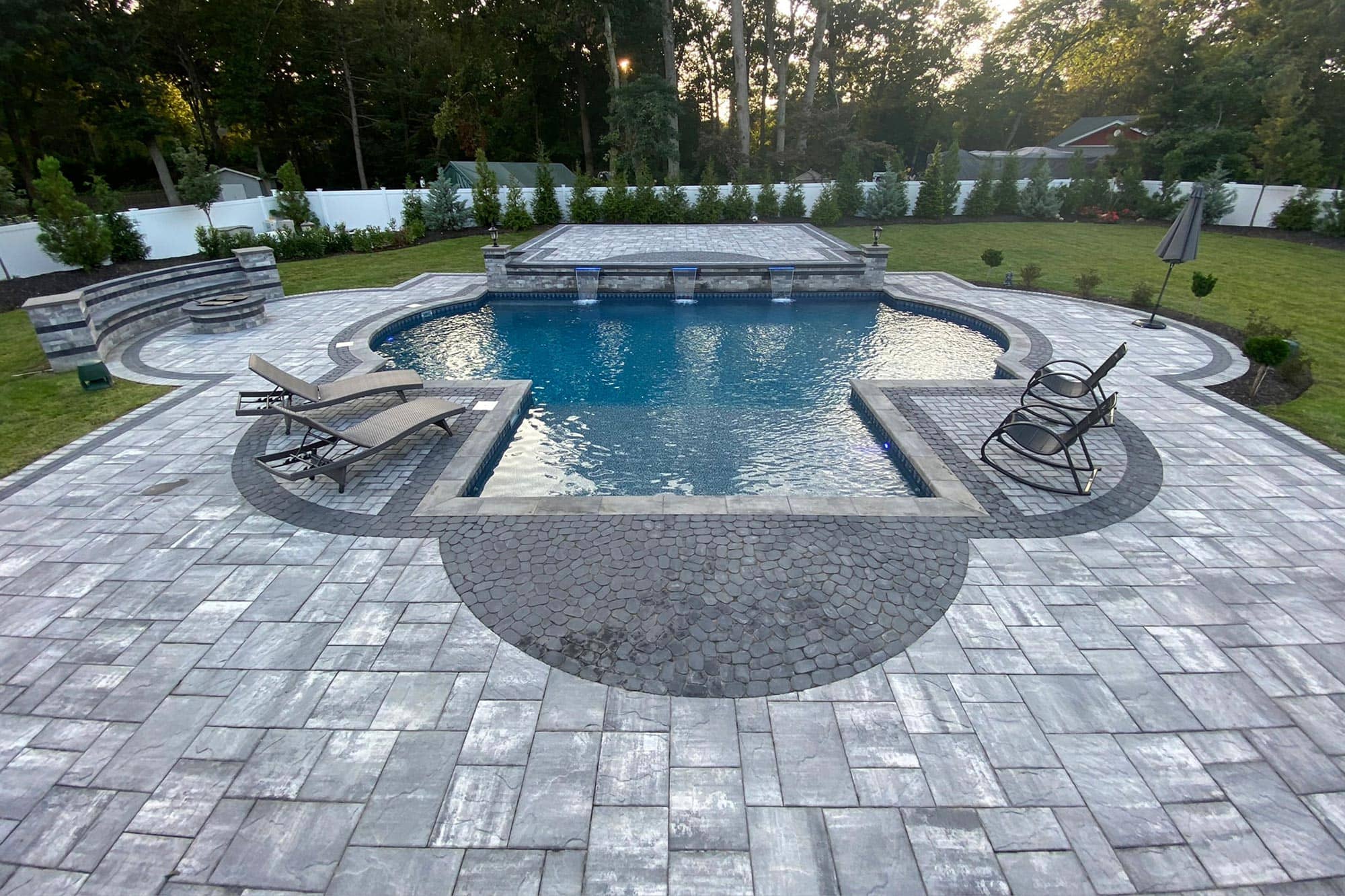A backyard with a rectangular swimming pool, poolside lounge chairs, stone patio, green lawn, and trees lining the fence in the background.
