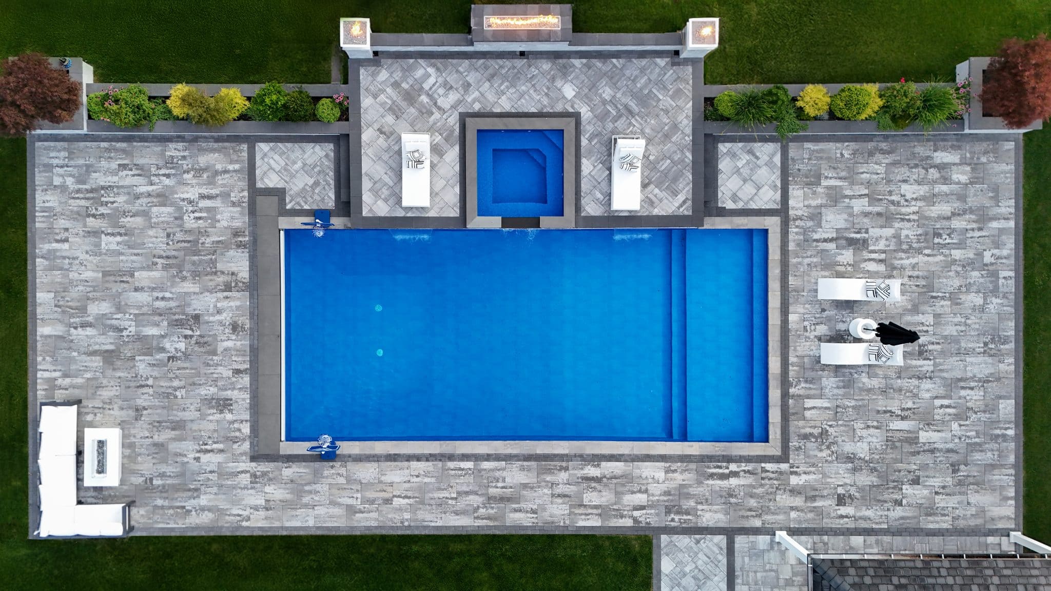 Aerial view of a rectangular New Vinyl Pool with a square hot tub, surrounded by stone tiles, lounge chairs, and landscaped greenery.