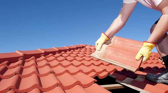 Ein Arbeiter in Handschuhen verlegt rote Dachziegel unter klarem blauem Himmel.