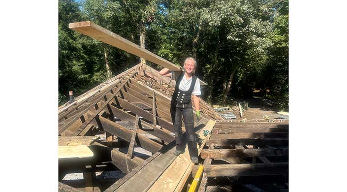 Eine Person steht auf einer Baustelle im Freien, trägt Arbeitskleidung und hält ein langes Holzbrett auf der Schulter, während sie lächelt.