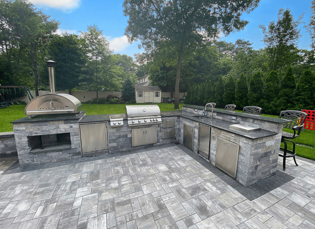 Outdoor kitchen with stainless steel appliances, including a grill, pizza oven, and sink, set in a stone counter. The area is surrounded by trees and a garden.
