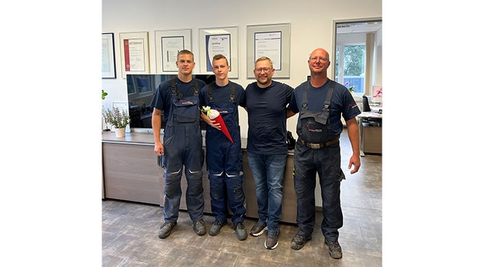 Drei Männer stehen nebeneinander, zwei tragen Arbeitskleidung, einer hält einen Blumenstrauß. Sie lächeln, im Hintergrund sind gerahmte Zertifikate an der Wand.