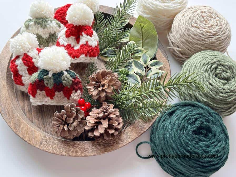 Wooden tray with small crocheted red, white, and green hats, pinecones, greenery, berries; 3 yarn balls sit nearby.Free crochet mini hat christmas ornament pattern