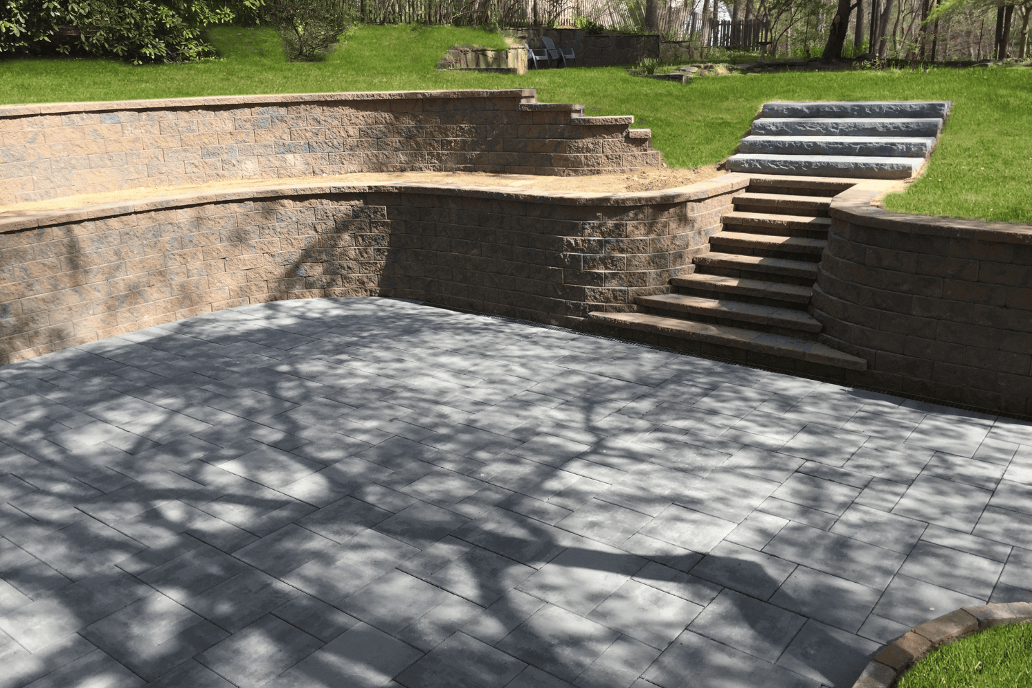 Tiered retaining walls with stone steps lead from a paved patio area up to a grassy lawn; tree shadows are cast on the patio surface.