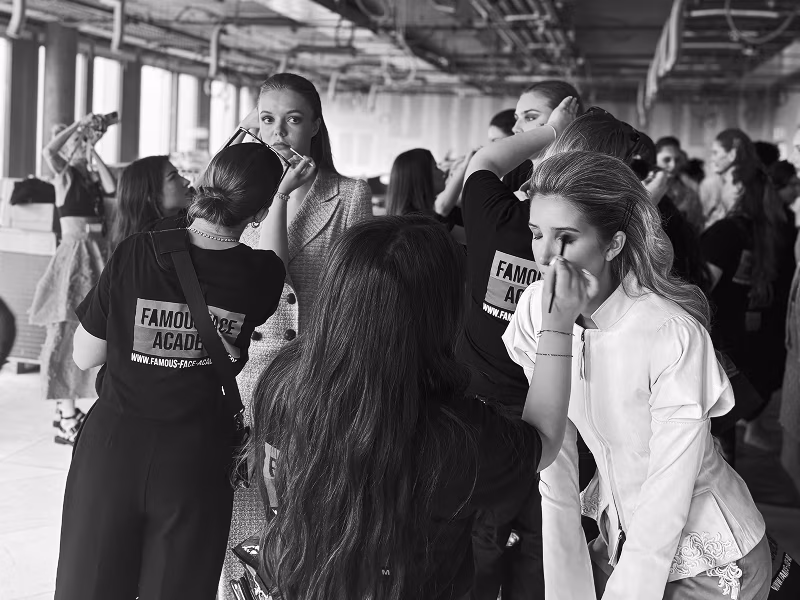 Mehrere Personen in einem großen Raum, zwei tragen T-Shirts mit der Aufschrift 'FAMOUS ACADEMY', eine Person trägt weiße Kleidung und wird geschminkt