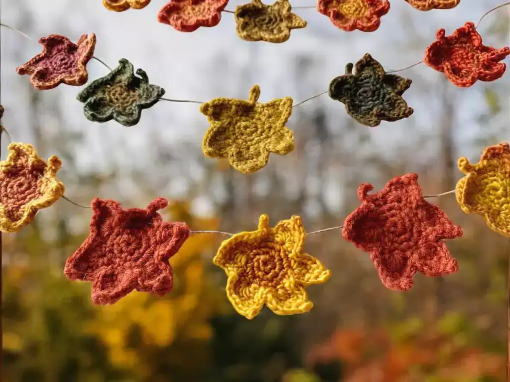 Crocheted autumn leaves, including maple designs in fall colors, hang on twine outdoors with blurred trees in the background. Free crochet pattern