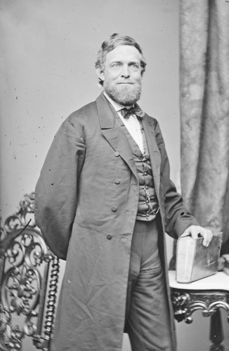 NPG.81.M71 Victorian man in formal suit with beard standing near ornate table.