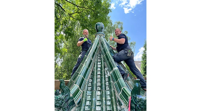Zwei Männer stehen auf einer gläsernen Dachkonstruktion, lächeln und zeigen Daumen hoch. Im Hintergrund sind grüne Bäume und ein blauer Himmel zu sehen.