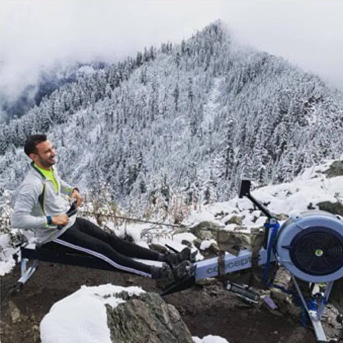 A man uses a rowing machine outdoors on a snowy mountain, with snow-covered trees and slopes in the background.
