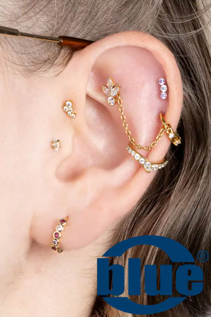 Close-up of a ear with multiple gold crystal earrings and a delicate chain cuff, plus a blue logo overlay in the corner.